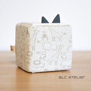 May include: A white fabric cube with a black felt ear-like detail on top. The cube is printed with a pattern of animals, including giraffes, elephants, and lions, in a light brown color.