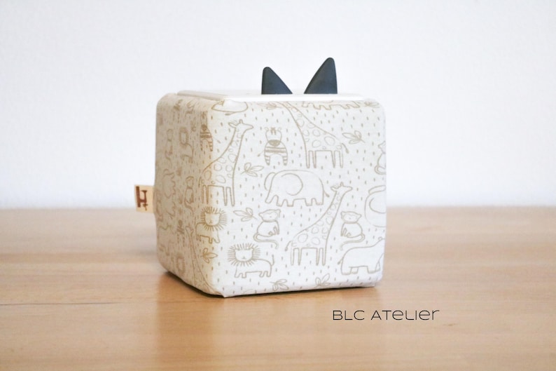 May include: A white fabric cube with a black felt ear-like detail on top. The cube is printed with a pattern of animals, including giraffes, elephants, and lions, in a light brown color.