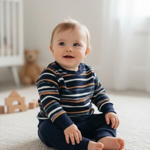 Könnte beinhalten: Ein Baby in einem marineblauen, gestreiften Langarmshirt und einer passenden Hose sitzt auf einem hellen Teppich. Das Shirt hat Streifen in Weiß, Braun und Blau. Im Hintergrund sind ein Kinderbett und ein Teddybär zu sehen.