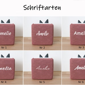 May include: Six pink fabric boxes with black cat ears on top. Each box has the name "Amelie" written on it in a different white font style. The boxes are numbered 1 through 6.