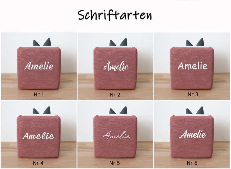 May include: Six pink fabric boxes with black cat ears on top. Each box has the name "Amelie" written on it in a different white font style. The boxes are numbered 1 through 6.