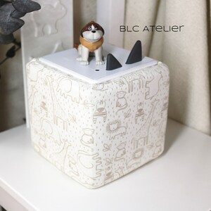 May include: A white cube-shaped speaker with a cream-colored fabric cover featuring a repeating pattern of animals, including giraffes, elephants, and lions. The speaker has two black triangular cutouts on the top and a small brown dog figurine on top of the speaker. The text "BLC ATELIER" is printed on the speaker.