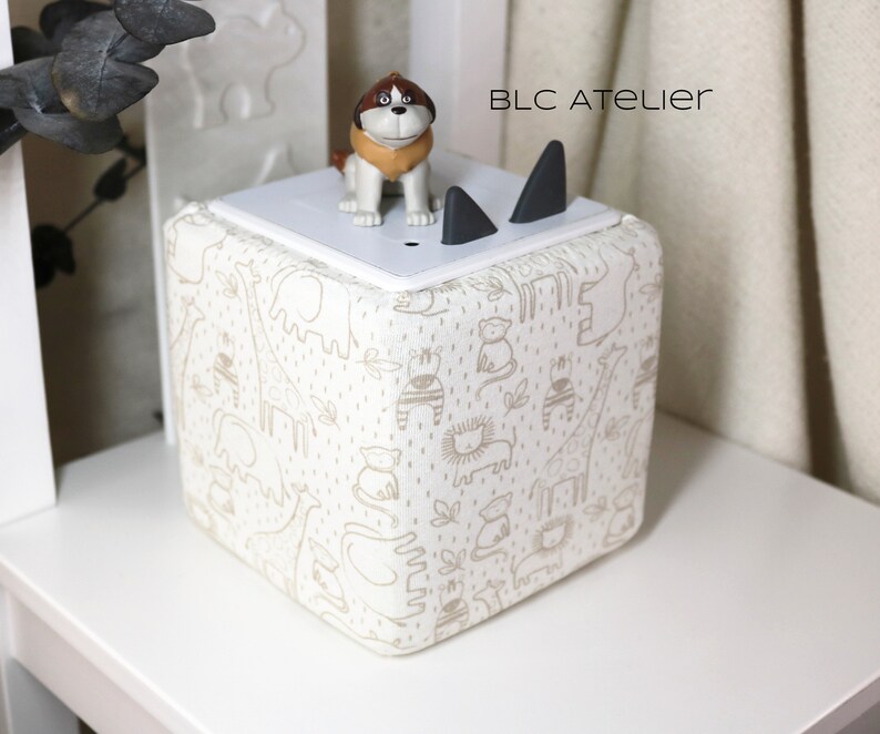May include: A white cube-shaped speaker with a cream-colored fabric cover featuring a repeating pattern of animals, including giraffes, elephants, and lions. The speaker has two black triangular cutouts on the top and a small brown dog figurine on top of the speaker. The text "BLC ATELIER" is printed on the speaker.