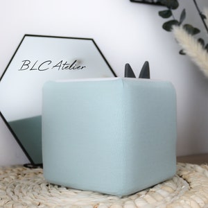 Könnte beinhalten: Ein hellblauer, würfelförmiger Hocker mit schwarzen Katzenohren oben. Der Hocker steht auf einem gewebten Teppich.