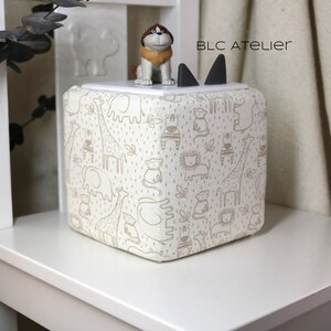 May include: A white fabric storage cube with a brown animal print design. The cube has a black cat-shaped lid with a brown dog figurine on top. The text "BLC ATELIER" is printed on the lid.