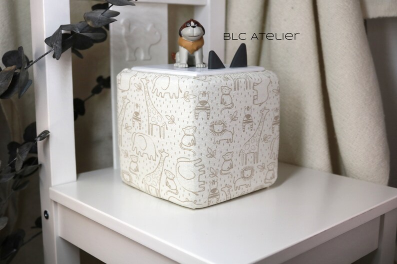 May include: A white fabric storage cube with a brown animal print design. The cube has a black cat-shaped lid with a brown dog figurine on top. The text "BLC ATELIER" is printed on the lid.