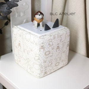 May include: A white cube-shaped storage box with a cream and brown animal print design. The box has a white lid with two black cat ears on top. A small brown and white dog figurine is sitting on the lid. The text "BLC ATELIER" is printed on the lid.