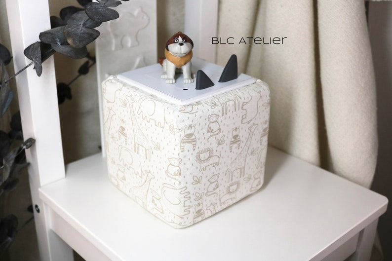 May include: A white cube-shaped storage box with a cream and brown animal print design. The box has a white lid with two black cat ears on top. A small brown and white dog figurine is sitting on the lid. The text "BLC ATELIER" is printed on the lid.