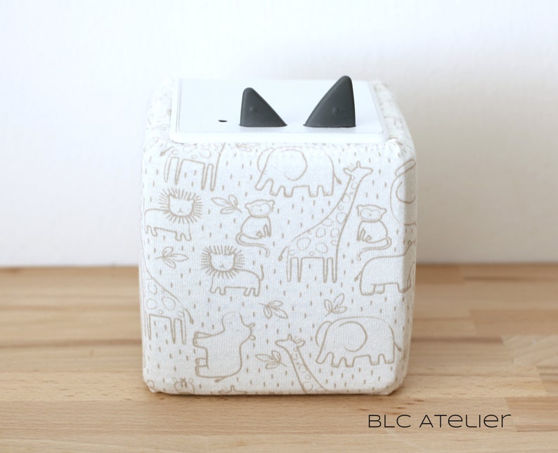 May include: A white cube-shaped storage box with a light brown fabric cover featuring a repeating pattern of animals, including elephants, giraffes, lions, and monkeys. The fabric cover has a subtle dotted texture. The box has two black ear-shaped handles on the top.