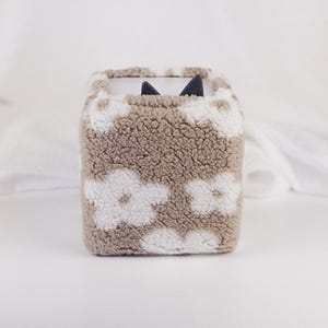 Könnte beinhalten: Ein quadratischer, beigefarbener Behälter mit einer strukturierten, flauschigen Außenseite. Weiße Blumenformen sind über die Oberfläche verstreut. Zwei schwarze Katzenohren schauen aus der oberen Öffnung. Der Behälter steht auf einer weißen Oberfläche.
