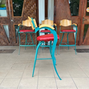 Può includere: Quattro sedie in metallo turchese con cuscini rossi e schienali in legno. Le sedie sono disposte in fila su un patio piastrellato.