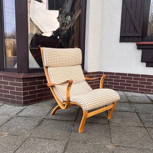 Puede incluir: Un sillón de madera con un cojín y respaldo a cuadros de color claro. La silla tiene brazos y patas de madera curvados. La silla está colocada en un patio de piedra gris. El fondo incluye una ventana y una pared blanca.