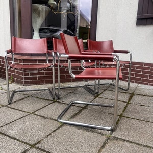 May include: Four vintage cantilever chairs with burgundy leather seats and backs. The chairs have a chrome-plated tubular steel frame. The chairs are arranged on a stone patio in front of a building.
