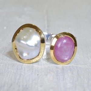 Op de afbeelding: Een ring met twee stenen met een grote, onregelmatige witte parel en een kleinere, ovale roze edelsteen. Beide stenen zijn gezet in gouden bezels, verbonden door een zilveren band. De ring ligt op een lichtgrijze ondergrond.