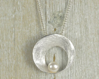 Puede incluir: Un collar de plata con un colgante curvo texturizado que contiene una perla blanca en el centro.