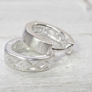 Pode incluir: Dois brincos de argola prateados. Cada brinco tem uma faixa grossa e arredondada com acabamento em metal escovado. A borda externa de cada brinco apresenta um design geométrico recortado em forma de diamante.