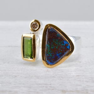 Könnte beinhalten: Ein Silberring mit einem großen, dreieckigen dunklen Opal mit blauem und grünem Schimmer, einem rechteckigen grünen Turmalin und einem kleinen runden braunen Edelstein. Jeder Stein ist in einer Goldfassung auf einer Silberschiene gefasst.