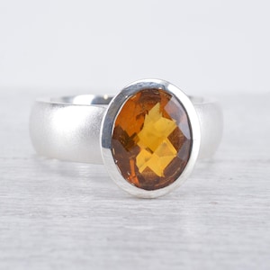 Puede incluir: Un anillo de plata con una gran piedra preciosa de color ámbar, de corte ovalado y facetado, engastada en un bisel liso y pulido. El cuerpo del anillo tiene un acabado mate y cepillado que contrasta con la superficie brillante de la piedra.