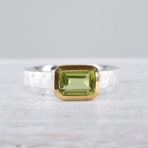 Könnte beinhalten: Ein Ring mit einem rechteckigen, hellgrünen Edelstein in einer Goldfassung. Die Ringschiene besteht aus Silber mit einer strukturierten, gebürsteten Oberfläche. Der Stein ist horizontal in der Mitte platziert und bildet einen Kontrast zwischen der goldenen Fassung und dem silbernen Ring.