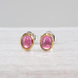 Puede incluir: Un par de pendientes de botón con piedras rosas translúcidas de forma ovalada en cabujón. Cada piedra está engastada en un bisel de oro amarillo pulido, montado sobre un poste de metal plateado con un cierre de mariposa estándar.