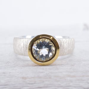 Könnte beinhalten: Ein silberner Ring mit einer strukturierten Band und einer goldenen Fassung mit einem klaren runden Edelstein.