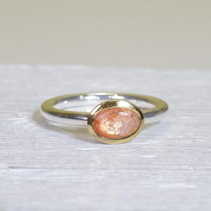 Könnte beinhalten: Ein silberner Ring mit einer goldenen Lünettenfassung, die einen ovalen, orangefarbenen Edelstein hält. Der Ring liegt auf einer hellen, strukturierten Oberfläche.