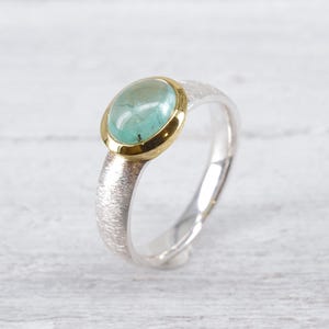 Könnte beinhalten: Ein Ring mit einem glatten, ovalen, hellgrünen Cabochon-Edelstein, der in einer polierten Gelbgoldfassung sitzt. Die Ringschiene besteht aus texturiertem Silber mit einer matten, gebürsteten Oberfläche, die einen Kontrast zwischen den Metallen bildet.