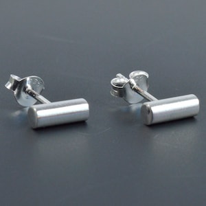 May include: A pair of silver bar stud earrings. Each earring features a small, cylindrical bar that is attached to a post with a butterfly backing.