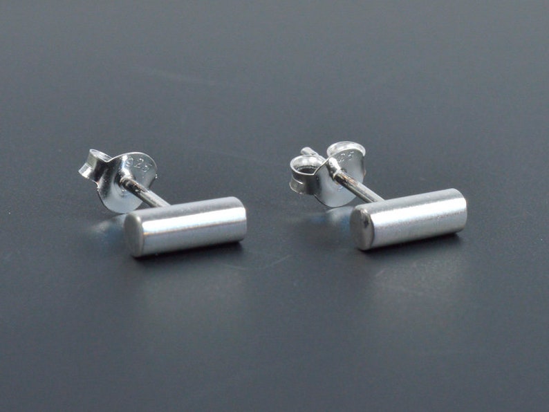 May include: A pair of silver bar stud earrings. Each earring features a small, cylindrical bar that is attached to a post with a butterfly backing.