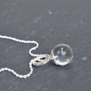 May include: A clear crystal ball pendant necklace with a silver chain. The pendant is a transparent sphere, attached to a silver loop. The chain is a twisted rope design. The necklace is displayed on a dark gray surface.
