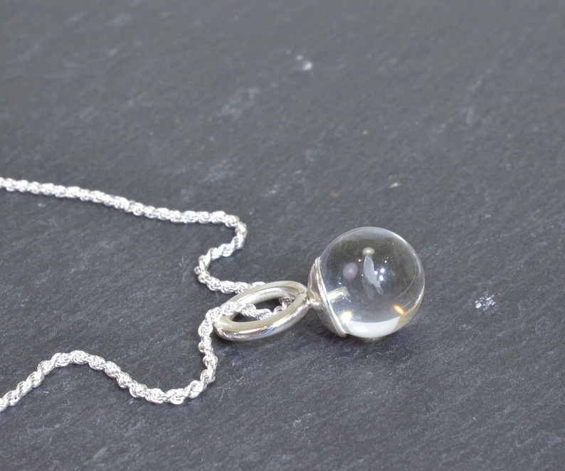 May include: A clear crystal ball pendant necklace with a silver chain. The pendant is a transparent sphere, attached to a silver loop. The chain is a twisted rope design. The necklace is displayed on a dark gray surface.