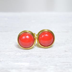 Puede incluir: Un par de pendientes de botón redondos con centros rojos brillantes engastados en metal dorado. Los pendientes tienen un diseño simple y clásico, con un acabado pulido. El color rojo es vibrante y llamativo.