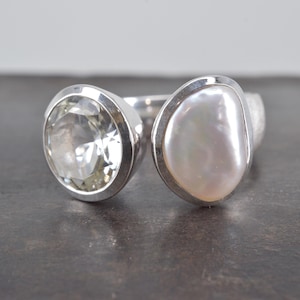 May include: A silver open-style ring featuring two distinct settings. One side holds a round, faceted clear gemstone, while the other side features an irregular, smooth, white iridescent pearl. The band is polished silver with a smooth finish.