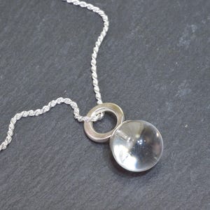 May include: A silver necklace featuring a clear, spherical pendant. The pendant is suspended from a silver ring and a twisted rope chain. The necklace is displayed against a dark gray background.