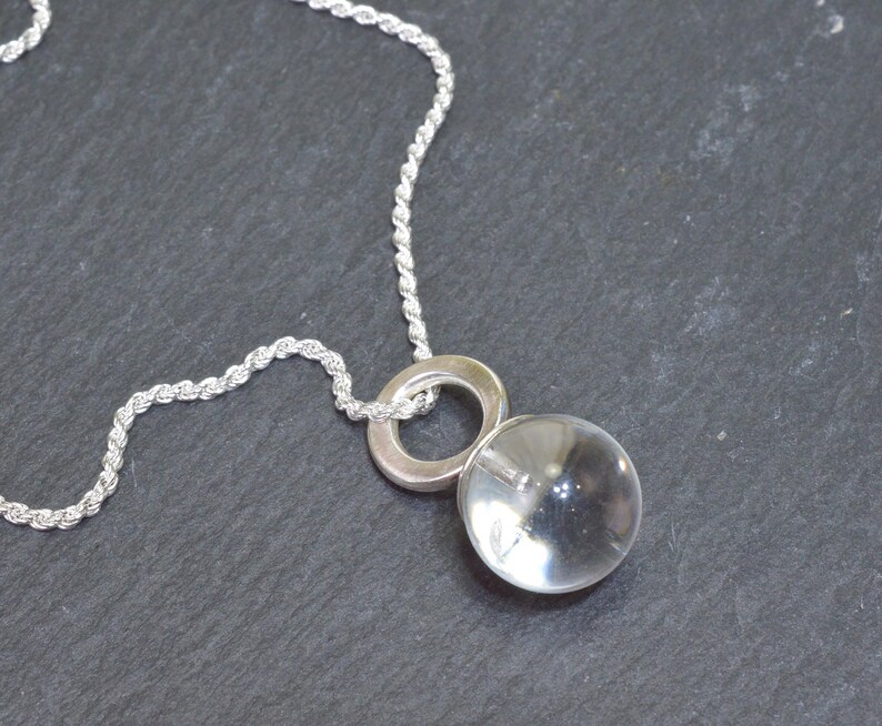 May include: A silver necklace featuring a clear, spherical pendant. The pendant is suspended from a silver ring and a twisted rope chain. The necklace is displayed against a dark gray background.