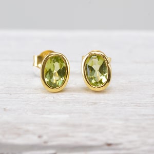 Könnte beinhalten: Ein Paar Ohrstecker mit oval geschliffenen, hellgrünen Peridot-Edelsteinen, die in polierte Gelbgoldfassungen eingefasst sind. Die Ohrringe haben ein klassisches Design mit einer glatten, metallischen Oberfläche, die jeden facettierten Stein umgibt.