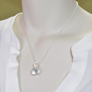 May include: A delicate silver necklace featuring a clear, round pendant. The pendant is suspended from a fine silver chain. The necklace is displayed on a white mannequin, against a soft, neutral background.