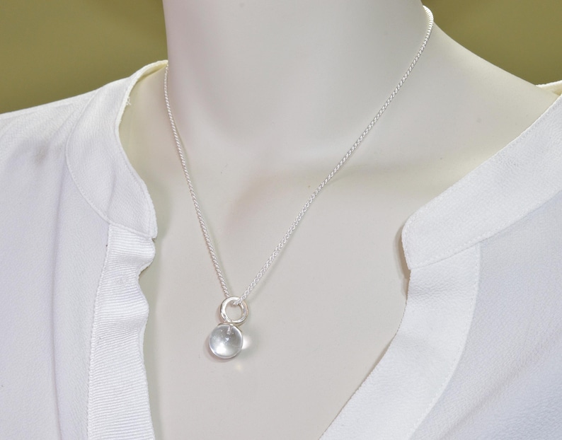 May include: A delicate silver necklace featuring a clear, round pendant. The pendant is suspended from a fine silver chain. The necklace is displayed on a white mannequin, against a soft, neutral background.