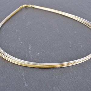 Könnte beinhalten: Eine mehrsträngige Halskette mit abwechselnd gold- und silberfarbenen Drähten. Die Halskette wird auf einer dunkelgrauen Oberfläche präsentiert. Der Verschluss ist goldfarben. Die Halskette ist ein Schmuckstück.