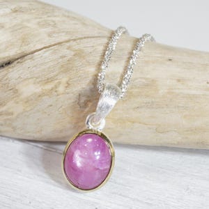 Könnte beinhalten: Eine silberne Halskette mit einem Anhänger mit einem ovalen, rosa Edelstein, der in einer goldfarbenen Lünette gefasst ist. Der Edelstein hat eine leuchtend rosa Farbe. Die Halskette liegt auf einem Stück Treibholz.