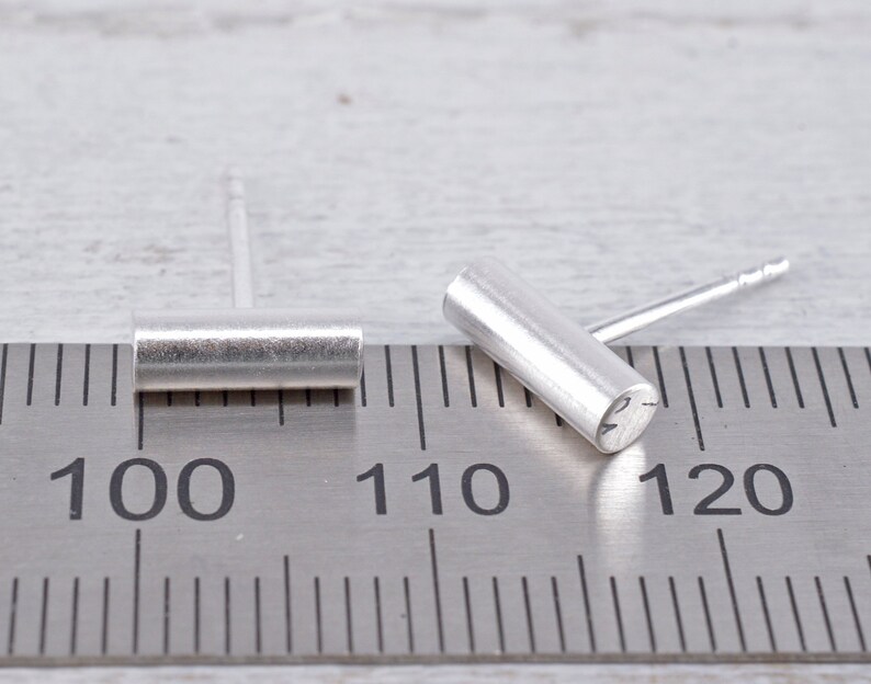 May include: Two silver bar earrings, one is laying flat on a ruler marked with inches, the other is standing upright. The ruler shows measurements from 100 to 120.