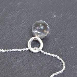 May include: A clear, spherical pendant necklace with a silver-colored ring and a rope chain. The pendant is a transparent orb, reflecting light. The necklace is displayed against a dark gray background, highlighting the jewelry's design.