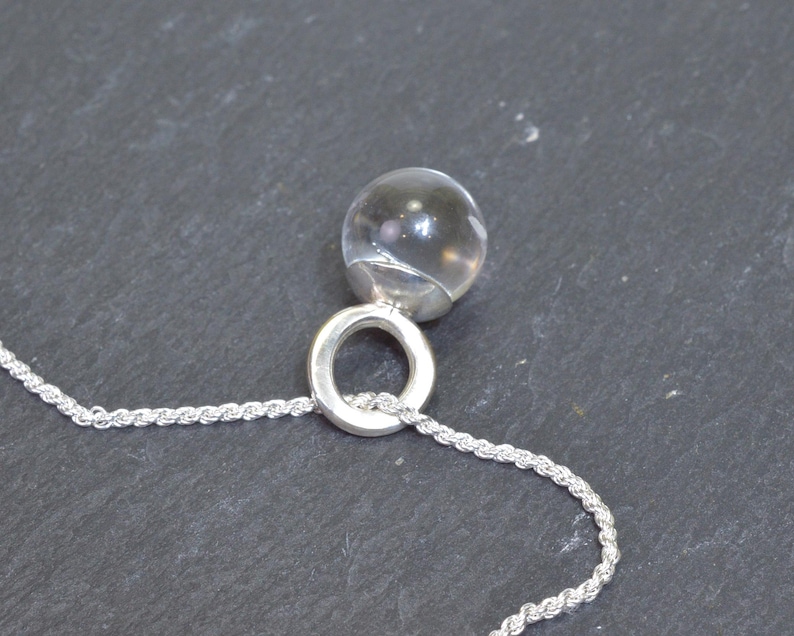 May include: A clear, spherical pendant necklace with a silver-colored ring and a rope chain. The pendant is a transparent orb, reflecting light. The necklace is displayed against a dark gray background, highlighting the jewelry's design.