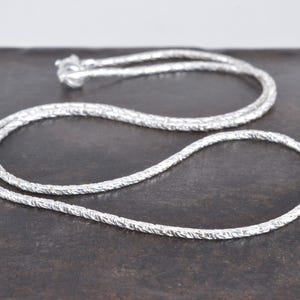 Könnte beinhalten: Eine silberfarbene Kettenhalskette mit einem texturierten, gedrehten seilartigen Design. Die Kette ist in einer kreisförmigen Schleife angeordnet und enthält einen kleinen Metallverschlussmechanismus, der oben sichtbar ist und ein einheitliches metallisches Finish aufweist.