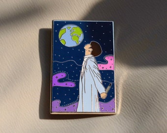 May include: Enamel pin featuring a person gazing at a globe in a starry night sky. The pin has a gold border and depicts a person in a white coat holding a knife. The background includes purple and pink cloud-like shapes.