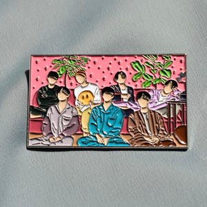 May include: Enamel pin featuring a group of people in pajamas sitting on a pink background with a dotted pattern. The pin has a silver border and includes green plant details.