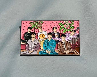 May include: Enamel pin featuring a group of people in pajamas sitting on a pink background with a dotted pattern. The pin has a silver border and includes green plant details.