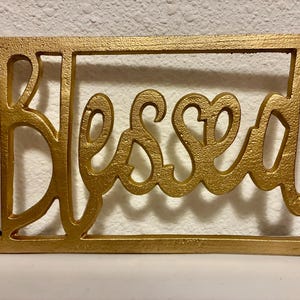 May include: A gold-colored, rectangular wall hanging with the word "Blessed" in a cursive font. The word is within a rectangular frame, creating a decorative piece for home decor.