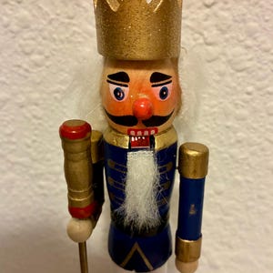 May include: A wooden nutcracker figurine in the shape of a king. The figurine has a gold crown, a red nose, and a white beard. It wears a blue jacket with gold accents and holds a staff. A classic holiday decoration.
