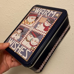 May include: A blue tin box with a red and white striped border. The box has a winter theme with snowmen and the words "Warm Wishes" and "Mittens for Sale".